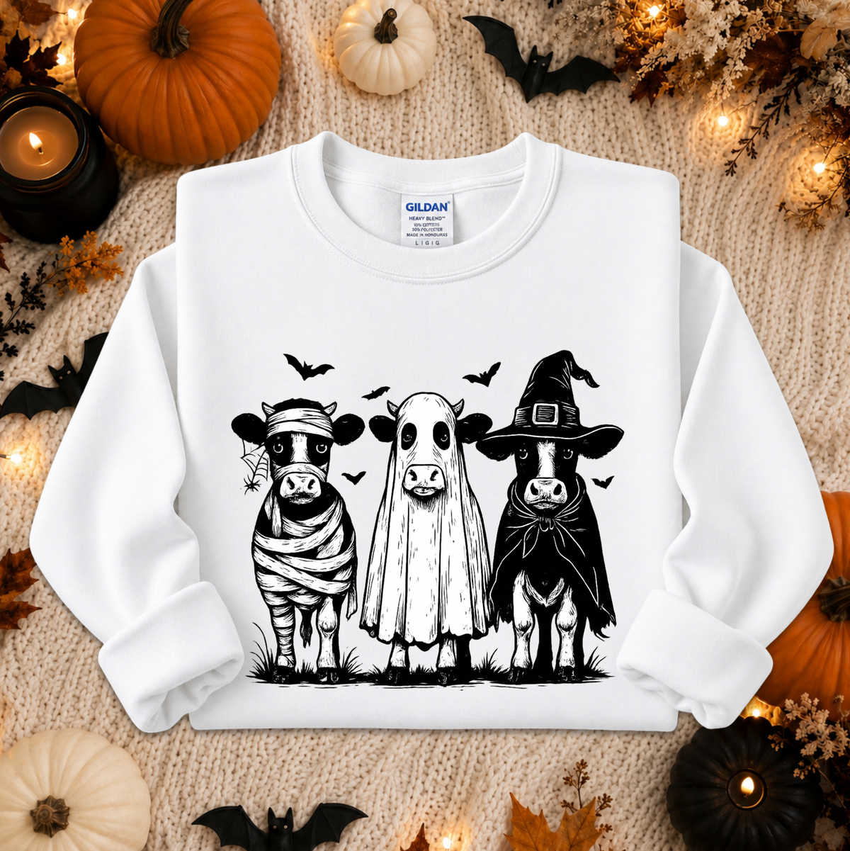 A white long-sleeve crewneck sweatshirt laid flat on a cozy beige knit blanket, surrounded by Halloween decorations including pumpkins, candles, leaves, and bats. The sweatshirt features a black graphic print of three cows dressed as a mummy, a ghost, and a witch.
