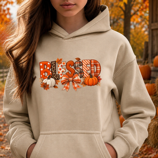 A woman wearing a beige hoodie with a 'Blessed' autumn graphic featuring pumpkins, leaves, and patterned letters, set against an outdoor fall background.