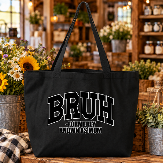 A black canvas tote bag with long handles sits on a rustic wooden table in a shop setting. The bag features the text 'BRUH -FORMERLY KNOWN AS MOM' in white varsity-style lettering.