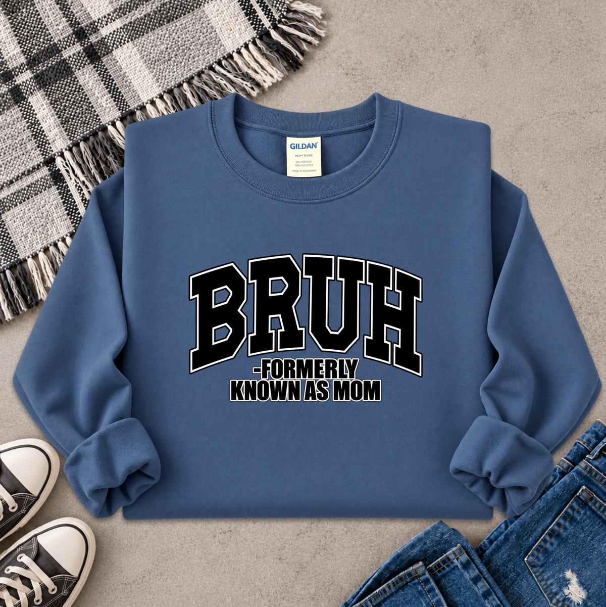 A blue crewneck sweatshirt with 'BRUH -FORMERLY KNOWN AS MOM' printed in black varsity-style lettering on the front. The sweatshirt is styled in a flat lay with a plaid throw blanket, black sneakers, and blue jeans on a gray concrete surface.