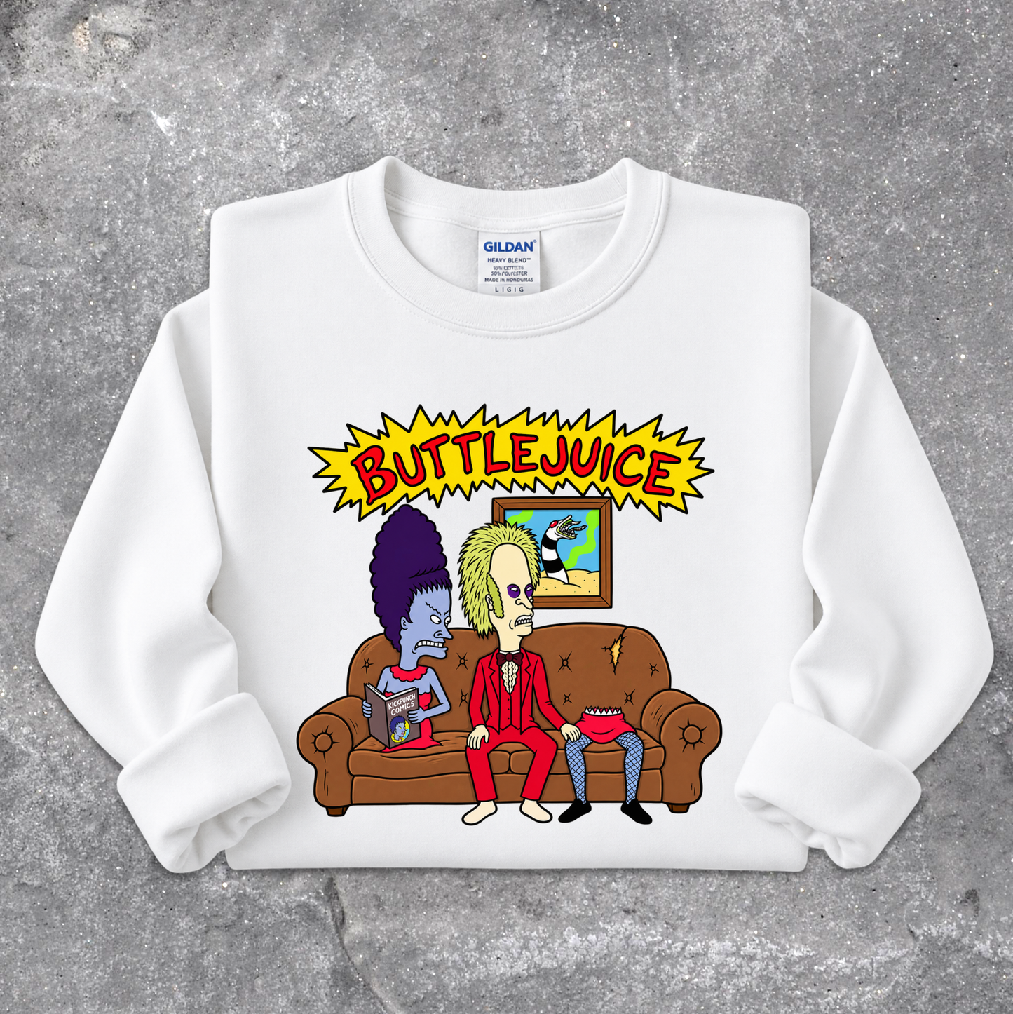 A white crewneck sweatshirt featuring a cartoon graphic of two characters sitting on a brown couch under a yellow 'BUTTLEJUICE' starburst graphic, laid flat on a textured gray concrete background. The neck label shows the brand Gildan.