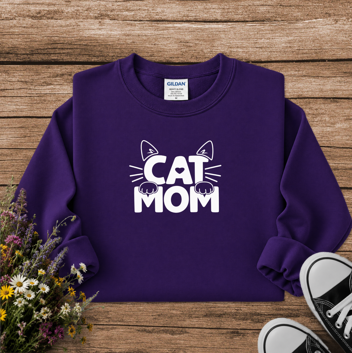 A purple crewneck sweatshirt with a white 'CAT MOM' graphic featuring cat ears, whiskers, and paws, laid flat on a rustic wooden background with flowers and sneakers as props. The neck label shows Gildan.