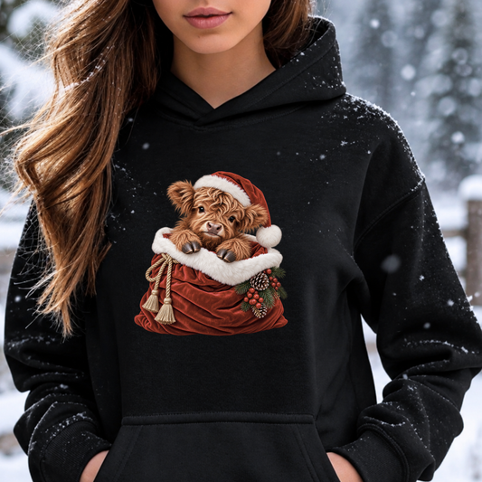 A woman wearing a black hoodie with a graphic of a fluffy brown cow calf in a red Santa hat and sack, photographed outdoors in the snow.