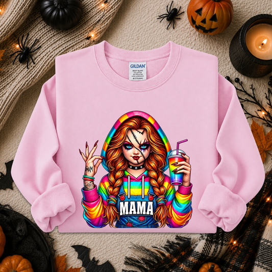 A light pink crewneck sweatshirt by Gildan, featuring a colorful graphic of a woman with long red-blonde braids holding a drink, with the word 'MAMA' printed below. The sweatshirt is displayed in a flat-lay arrangement with Halloween decorations including a pumpkin, spider, candles, and a knit sweater.