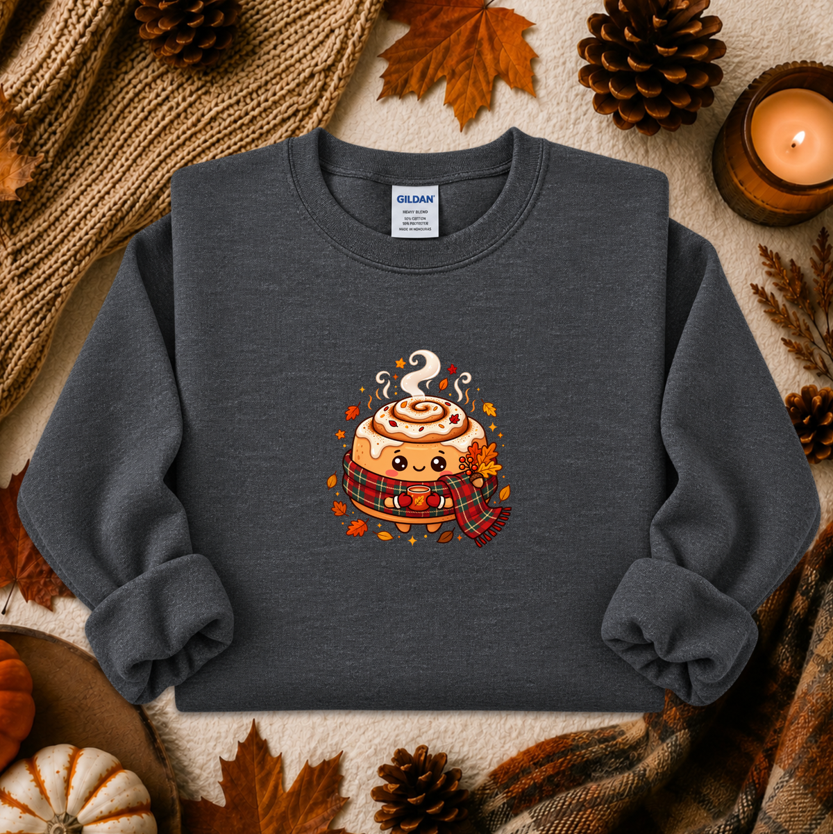 A dark gray crewneck sweatshirt featuring a cute cartoon cinnamon roll graphic with autumn accents, styled in a flat lay with fall-themed props like pinecones, a candle, and a knit blanket.