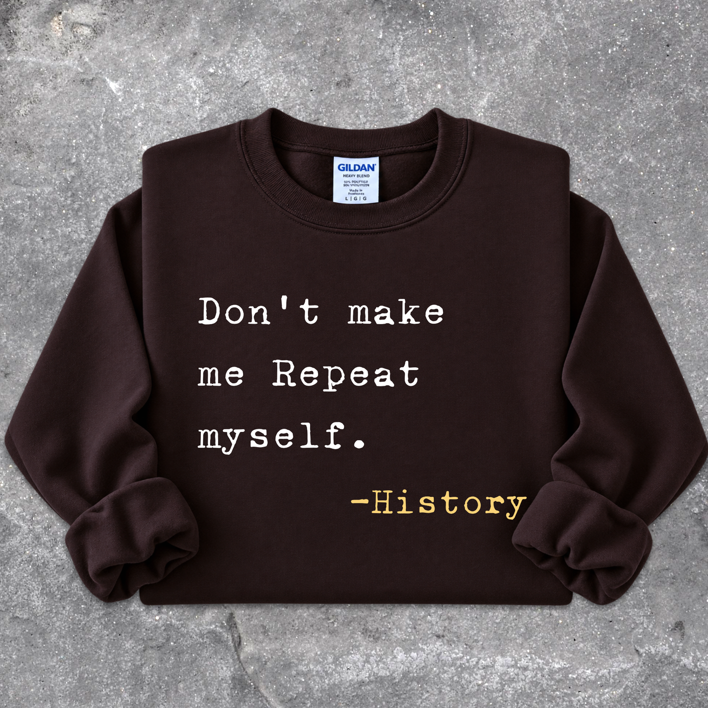 A dark brown crewneck sweatshirt with rolled cuffs, featuring the printed quote 'Don't make me Repeat myself. -History' in white and yellow text, laid flat on a textured gray concrete background.