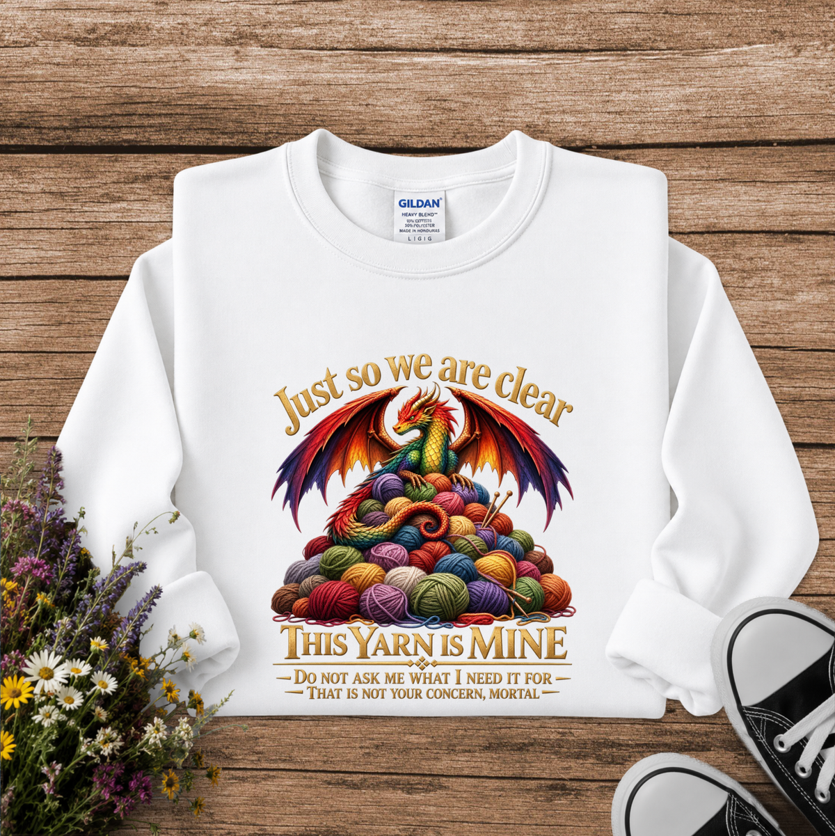 A white Gildan sweatshirt laid flat on a rustic wooden surface, decorated with a colorful graphic of a dragon rising from a pile of yarn balls, with the text 'Just so we are clear THIS YARN IS MINE'. Daisies and other wildflowers are on the left, and black and white sneakers are visible on the bottom right.
