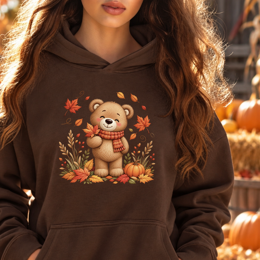 A woman wearing a dark brown hoodie with a graphic of a cute cartoon bear surrounded by autumn leaves, pumpkins, and a red scarf. The background is a warm, blurred fall harvest scene with pumpkins.