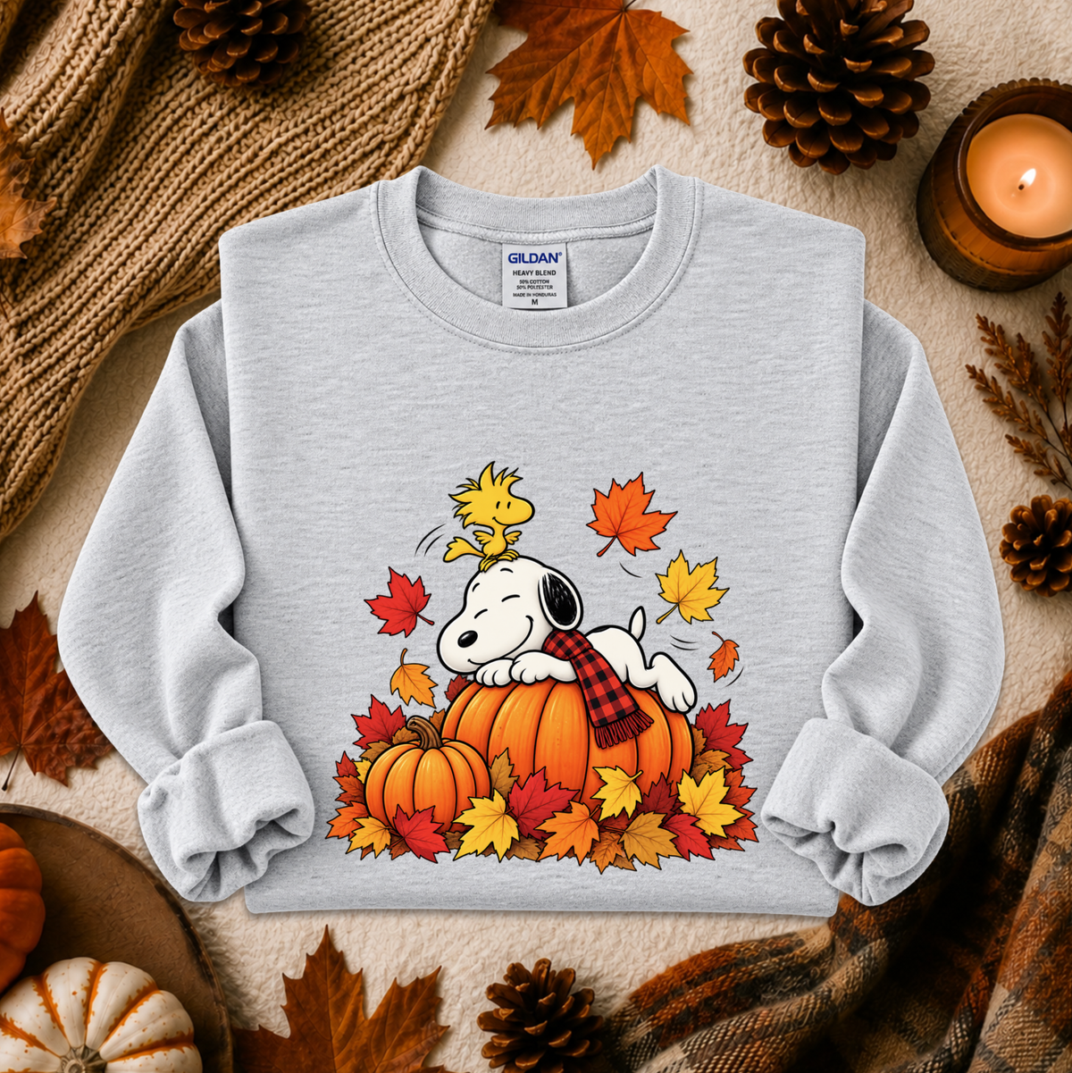 A light gray crewneck sweatshirt featuring a colorful graphic of Snoopy resting on a pumpkin surrounded by autumn leaves and Woodstock, styled in a cozy fall flat-lay with pinecones, a candle, and a knit blanket.