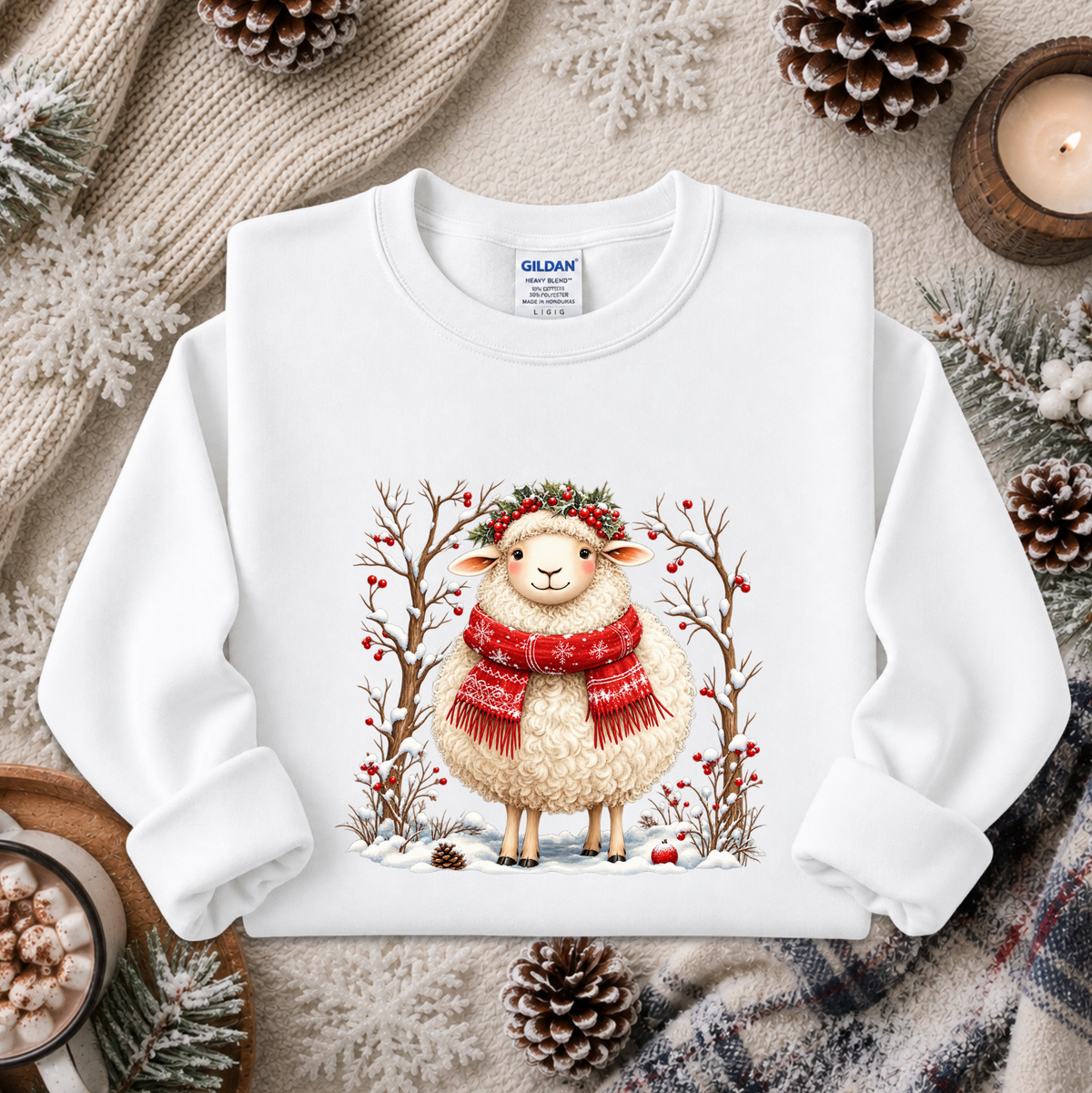 A white long-sleeve crewneck sweatshirt featuring a festive graphic of a smiling sheep in a red knit scarf and green wreath, surrounded by winter trees with red berries. The sweatshirt is styled in a cozy flat-lay arrangement with a knitted sweater, pinecones, a candle, and a mug of hot cocoa.