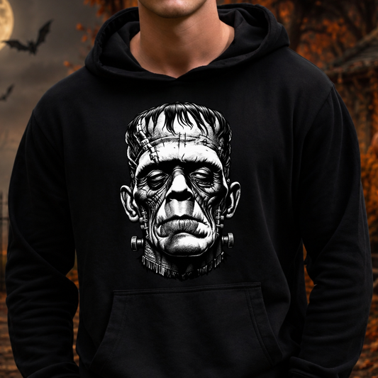 A person wearing a black pullover hoodie with a large black-and-white graphic of Frankenstein's monster face on the chest, set against a dark, spooky outdoor background with bats and autumn leaves.