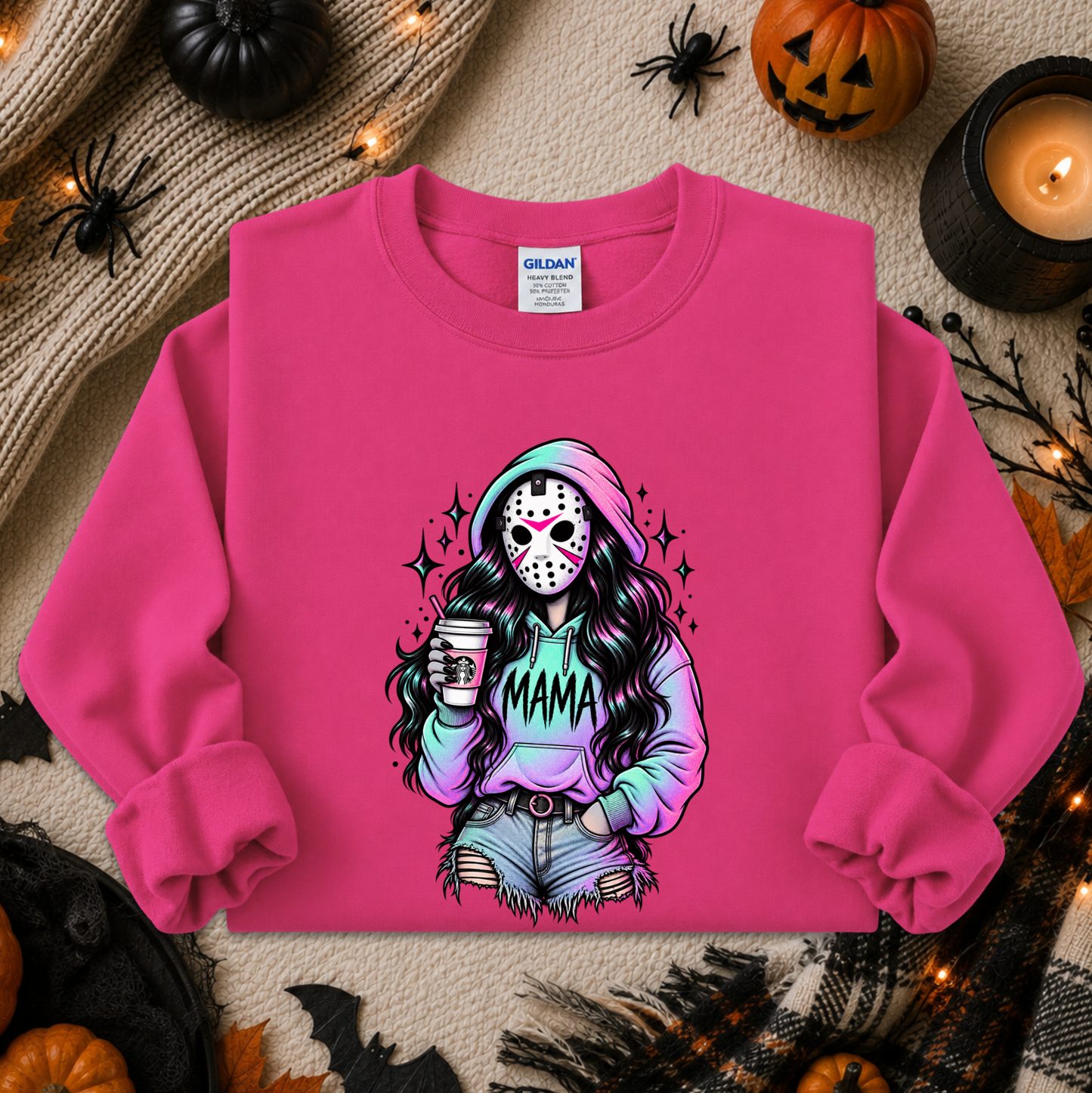 A bright pink crewneck sweatshirt laid flat on a cozy autumn-themed background. The sweatshirt features a colorful graphic of a woman in a hockey mask holding a coffee cup with 'MAMA' written across her hoodie.