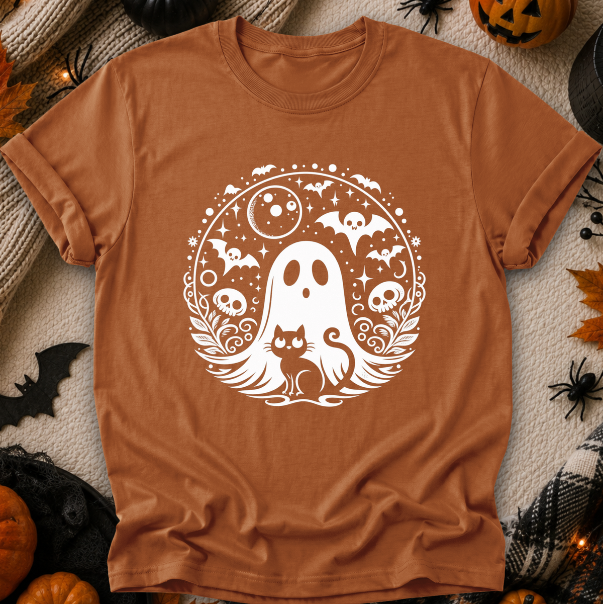 A rust-orange crewneck t-shirt with a white Halloween graphic of a ghost, cat, bats, and skulls, displayed flat on a cozy autumn-themed background with pumpkins, spider decorations, and a knit blanket.