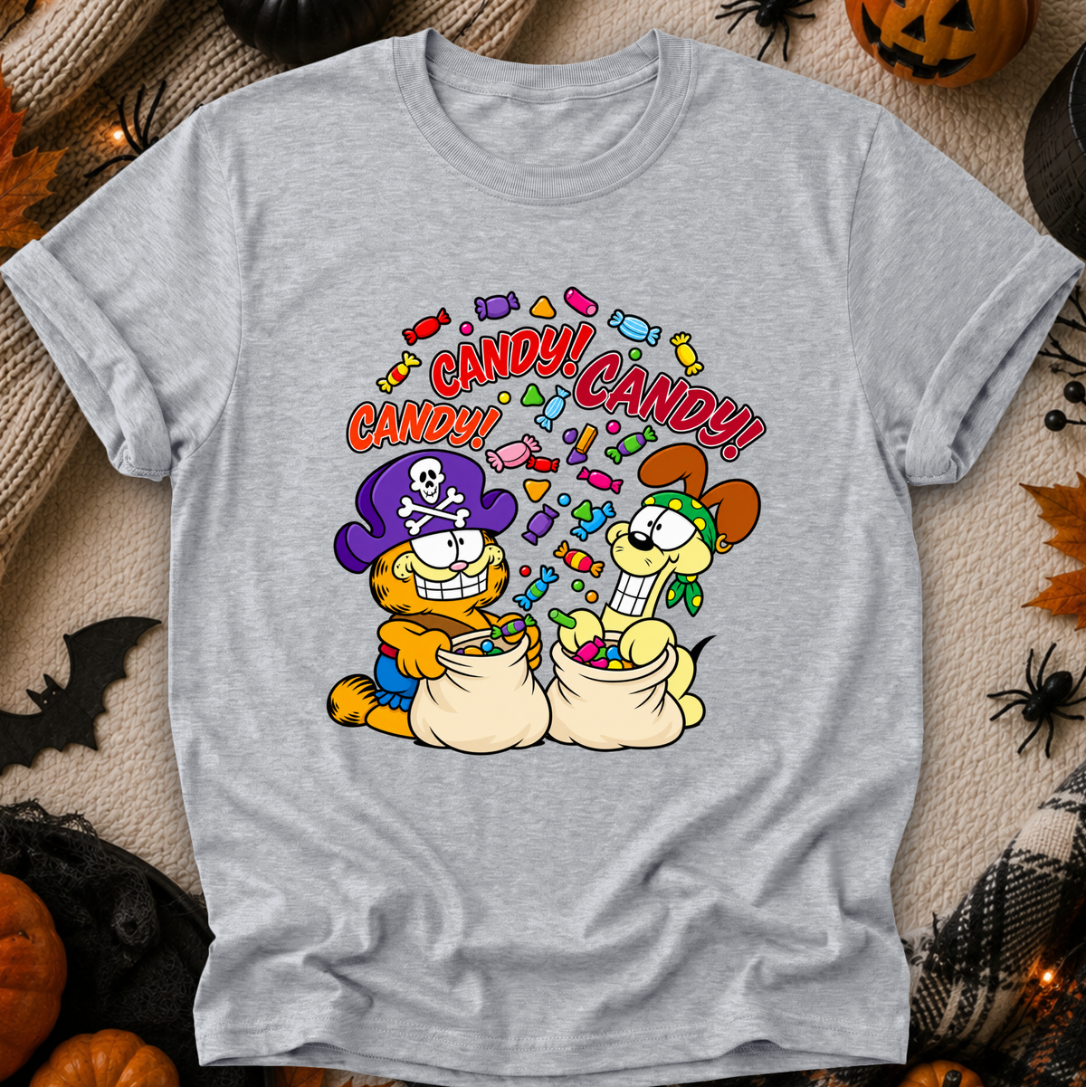 A light heather gray short-sleeve t-shirt with a colorful cartoon graphic of two dogs dressed as pirates and cats, surrounded by candy and the text 'CANDY! CANDY!'. The shirt is displayed flat on a cozy beige fabric background decorated with Halloween props like pumpkins, a spider, and a bat.