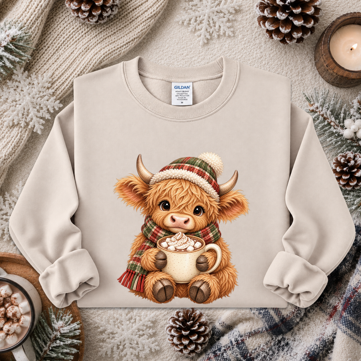 A beige crewneck sweatshirt featuring a cute cartoon Highland cow wearing a red and green plaid hat and scarf while holding a mug of hot cocoa with marshmallows. The sweatshirt is styled in a cozy winter flat-lay with pinecones, a candle, and knitted blankets.