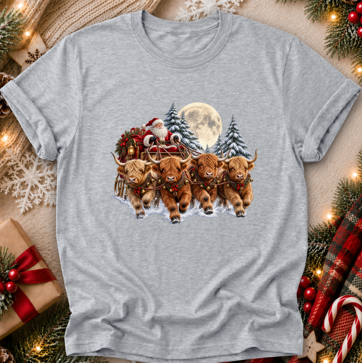 A heather gray short-sleeve t-shirt with a Christmas graphic print of four Highland cows pulling Santa's sleigh in the snow, surrounded by festive decorations like pine branches, lights, and a gift box.