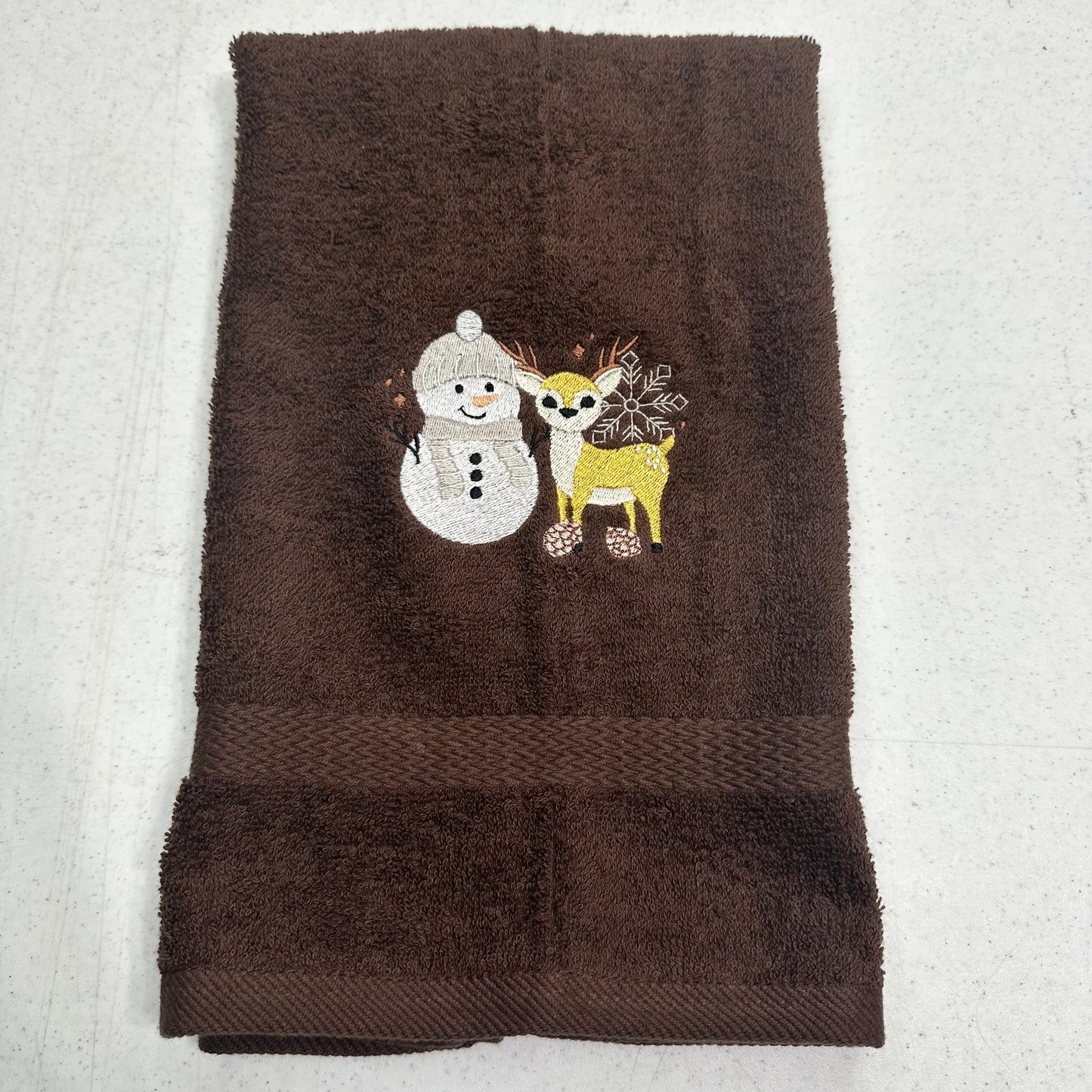 A dark brown terry cloth hand towel featuring an embroidered design of a white snowman and a yellow deer with antlers on the front, laid flat on a light gray textured surface.