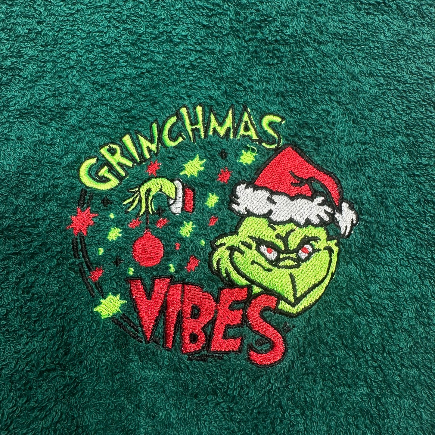 A close-up of a dark green textured fabric featuring an embroidered Grinch graphic with the words 'GRINCHMAS VIBES' and a Christmas ornament.