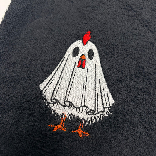 A close-up of a black textured fabric featuring an embroidered cartoon ghost chicken. The ghost has a white body, black eyes, a red comb, and orange feet and beak details.