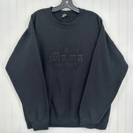 A black long-sleeve crewneck sweatshirt hanging on a black hanger against a white paneled wall. The front features an embossed tonal graphic reading 'Goth Mama Est.
