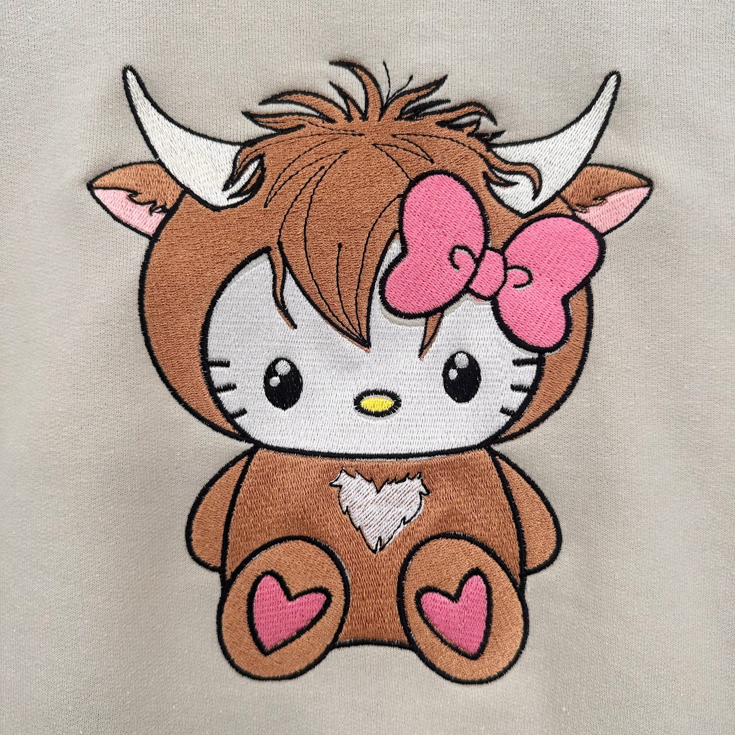 Embroidered design of hello kitty in a highland cow costume with a pink bow on a beige background