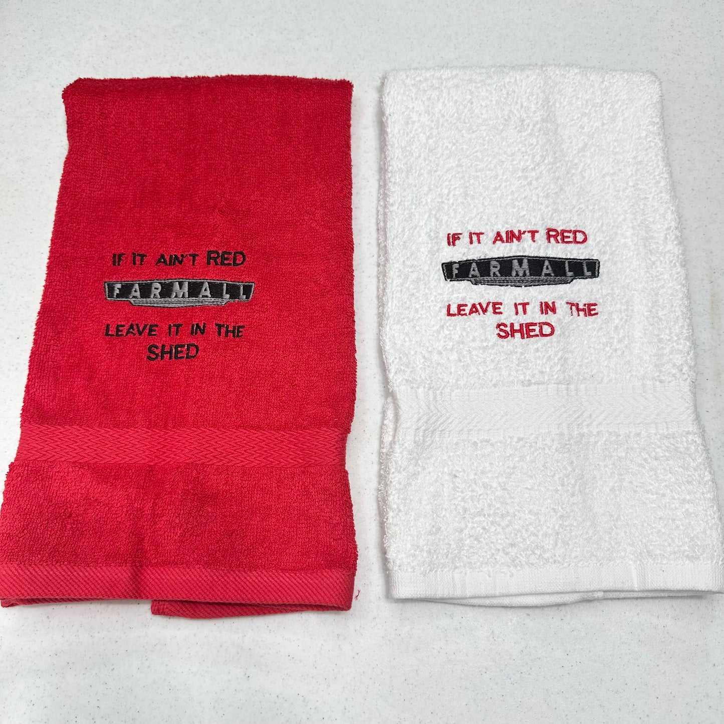 Two folded towels on a light gray background: one red towel with black embroidered text reading 'IF IT AIN'T RED FARMALL LEAVE IT IN THE SHED' and one white towel with red embroidered text reading 'IF IT AIN'T RED FARMALL LEAVE IT IN THE SHED'.