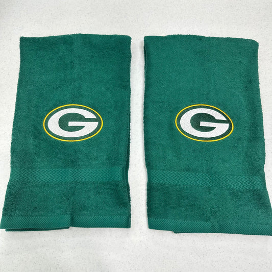 Two green towels with the Green Bay Packers 'G' logo, displayed side-by-side on a light gray textured background.