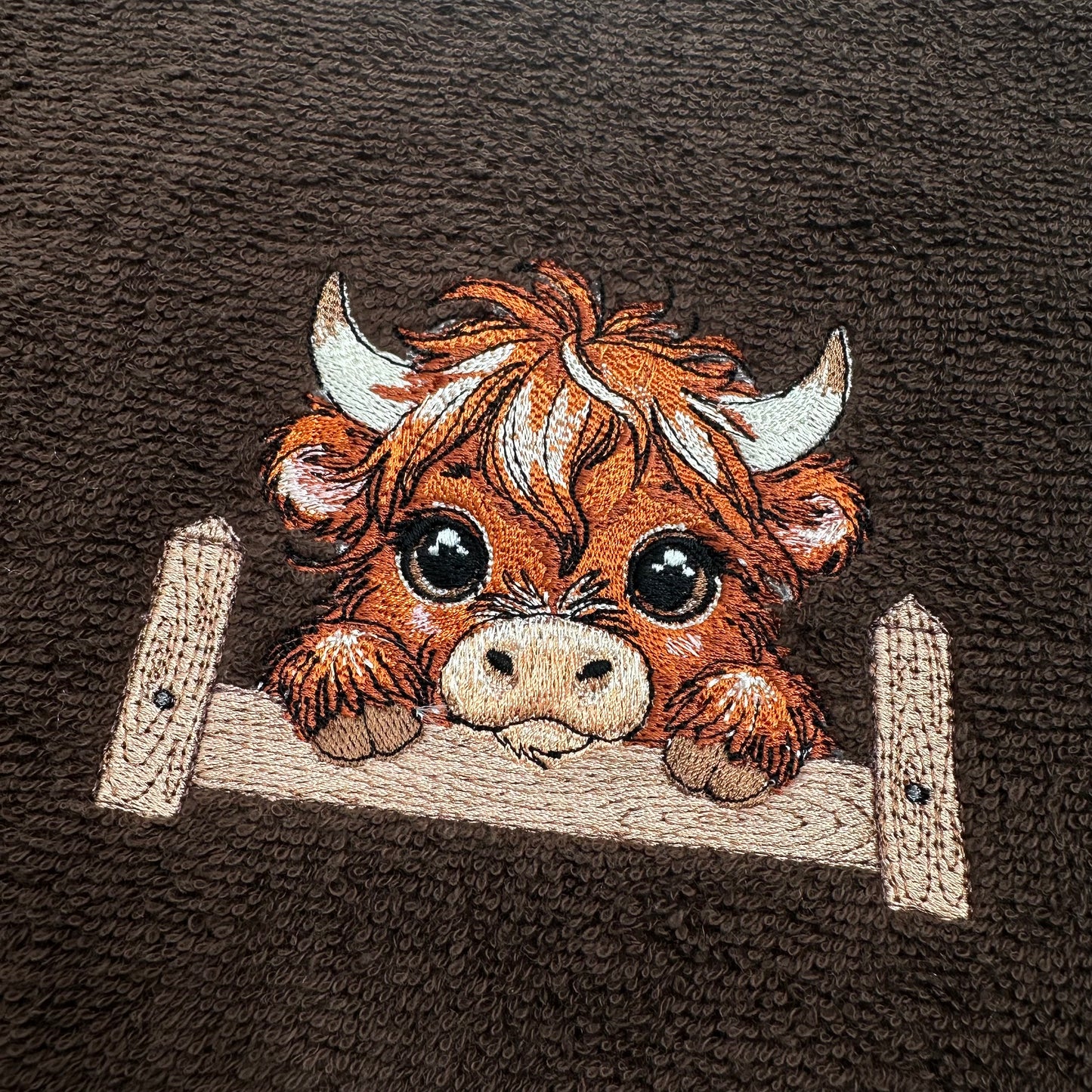 A close-up of a brown textured fabric featuring an embroidered cartoon Highland cow peeking over a light beige wooden fence. The cow has orange-brown hair, white horns, and large black eyes.