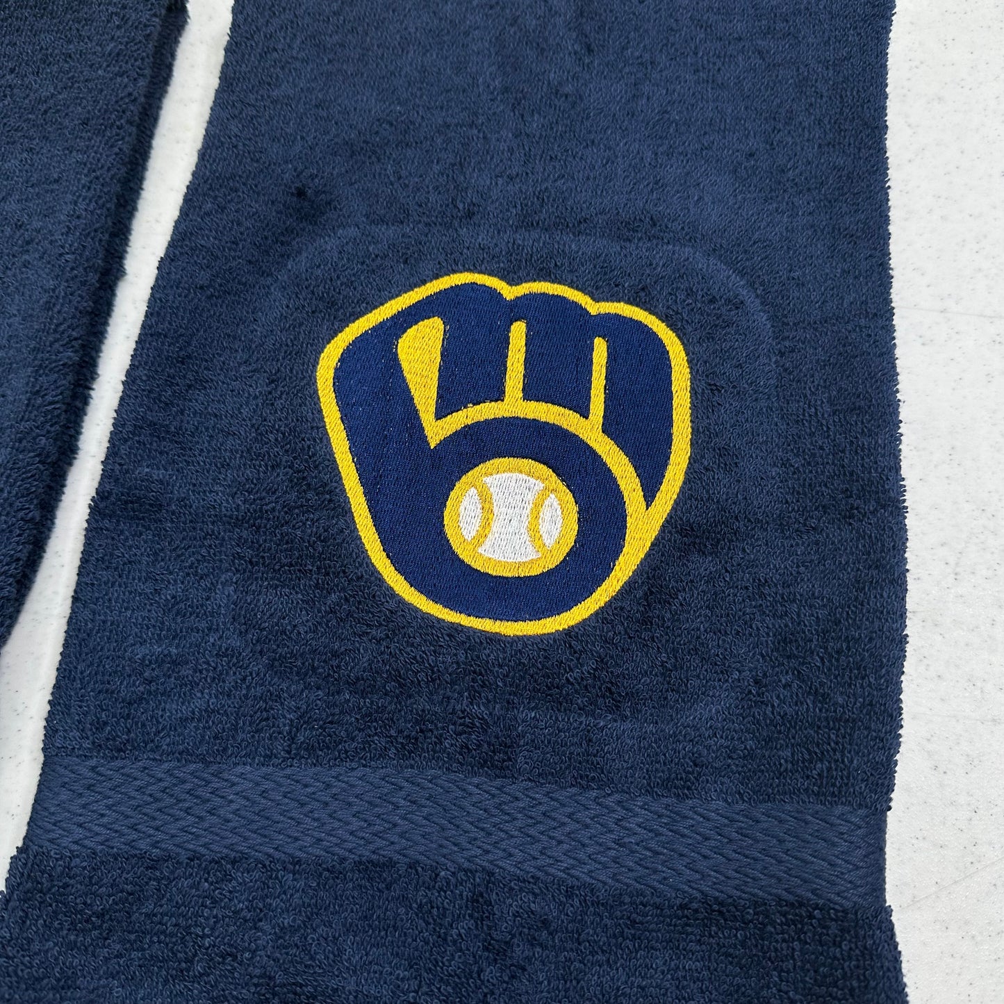 A close-up of a navy blue towel featuring the Milwaukee Brewers baseball glove and ball logo embroidered in yellow and white, with a matching navy woven band near the bottom.