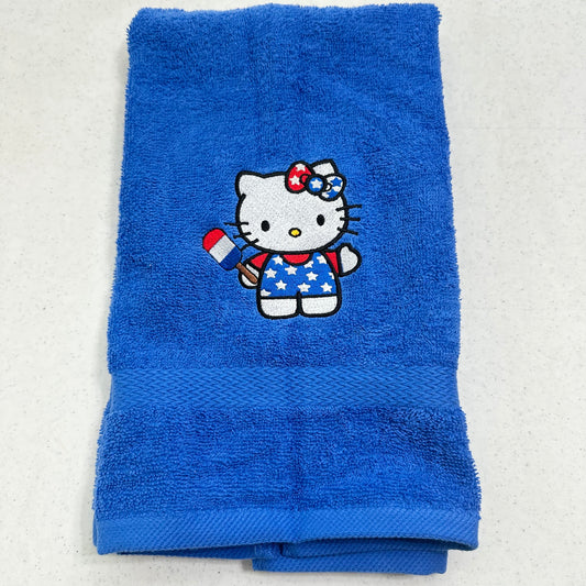 A folded blue towel featuring an embroidered Hello Kitty character holding a red, white, and blue flag, set against a light gray background.