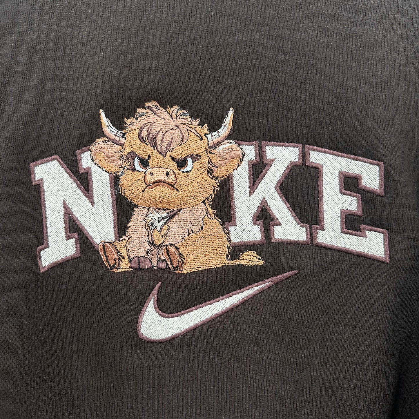 Close-up of a black sweatshirt featuring a vintage-style embroidered graphic with the word 'NIKE' in white varsity letters, a cartoon Highland cow mascot, and a white swoosh.