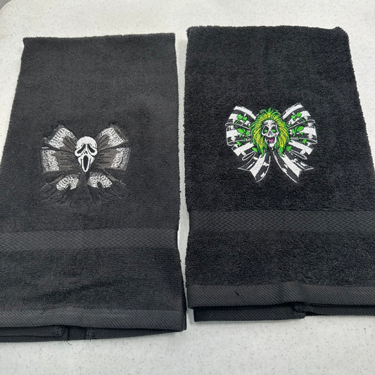 Two black towels laid flat on a light gray surface, each featuring a stitched horror-themed skull and ribbon motif. The towel on the left has a dark gray ghostface design, and the towel on the right has a green-and-white beetlejuice skull design.