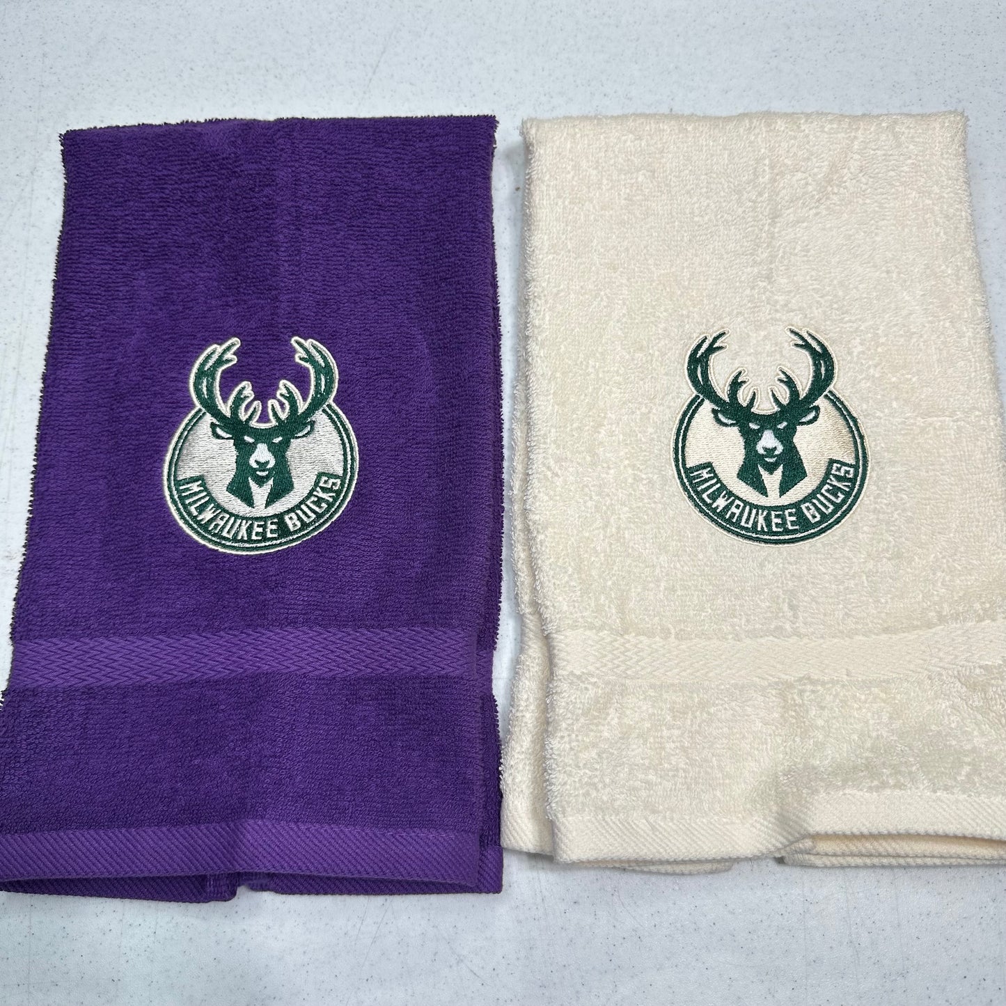 Two folded towels side by side on a light gray surface: one purple towel and one cream towel, each printed with a green Milwaukee Bucks deer logo and the words MILWAUKEE BUCKS.