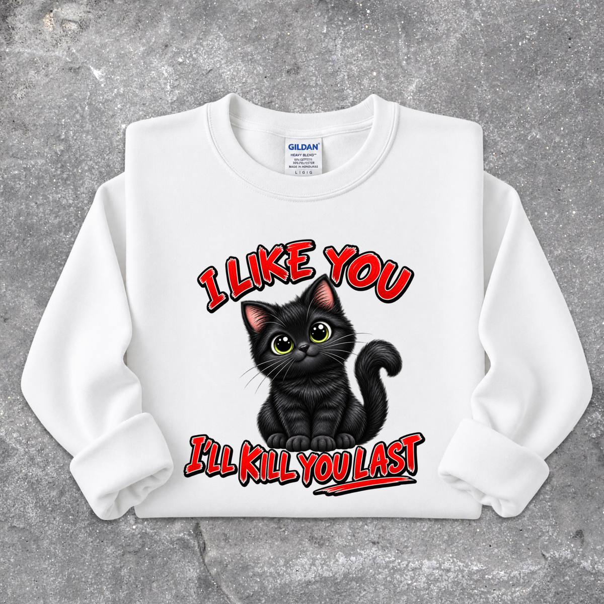 A white long-sleeve crewneck sweatshirt with a graphic of a black cat and the text 'I LIKE YOU I'LL KILL YOU LAST' in red and black, displayed flat on a gray concrete-textured background. The neck label shows Gildan.