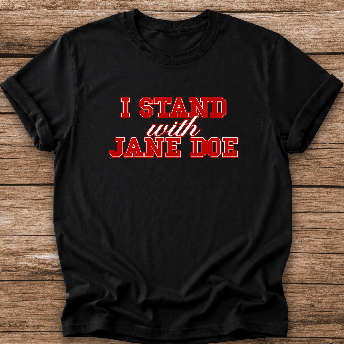 A black short-sleeve t-shirt with the slogan 'I STAND with JANE DOE' printed in red across the chest, laid flat on a rustic brown wooden plank background.