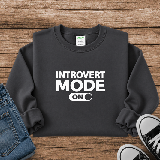A dark charcoal gray sweatshirt with 'INTROVERT MODE ON' printed in bold white text, styled in a flat lay on a rustic wooden background with blue jeans and black-and-white sneakers.
