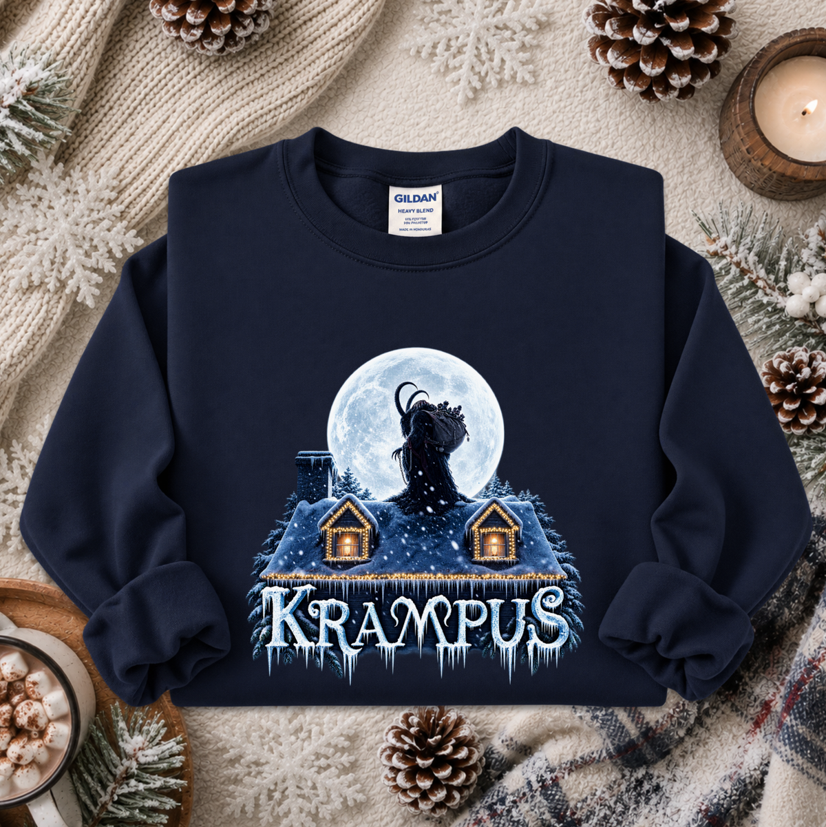A navy blue crewneck sweatshirt with a graphic of a goat-like creature in front of a full moon above two small houses, with the word 'KRAAMPUS' in icy letters below. The sweatshirt is displayed in a flat-lay arrangement with a cozy winter background including pinecones, a candle, and a knit blanket.