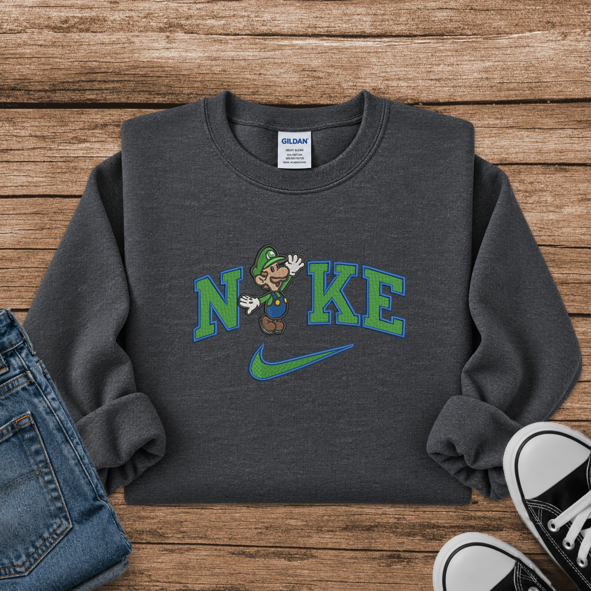 A dark gray crewneck sweatshirt featuring a graphic with Luigi and a Nike swoosh, styled in a flat lay on a wooden background with jeans and sneakers.