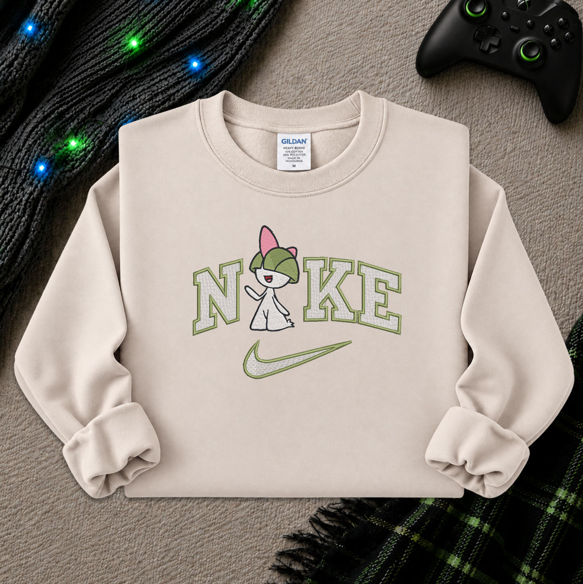 A beige crewneck sweatshirt featuring a graphic of a pokemon Raltz cartoon character wearing a green cap and holding a sign that says 'NO KE' with a green Nike swoosh underneath. The sweatshirt is styled flat on a gray textured surface with a black knit sweater, colorful fairy lights, and a black game controller in the background.