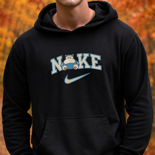 A person wearing a black hoodie with a light blue and white 'N'IKE' graphic across the chest, featuring a cartoon pokemon snorlax and a Nike swoosh, set against a blurred autumn background.