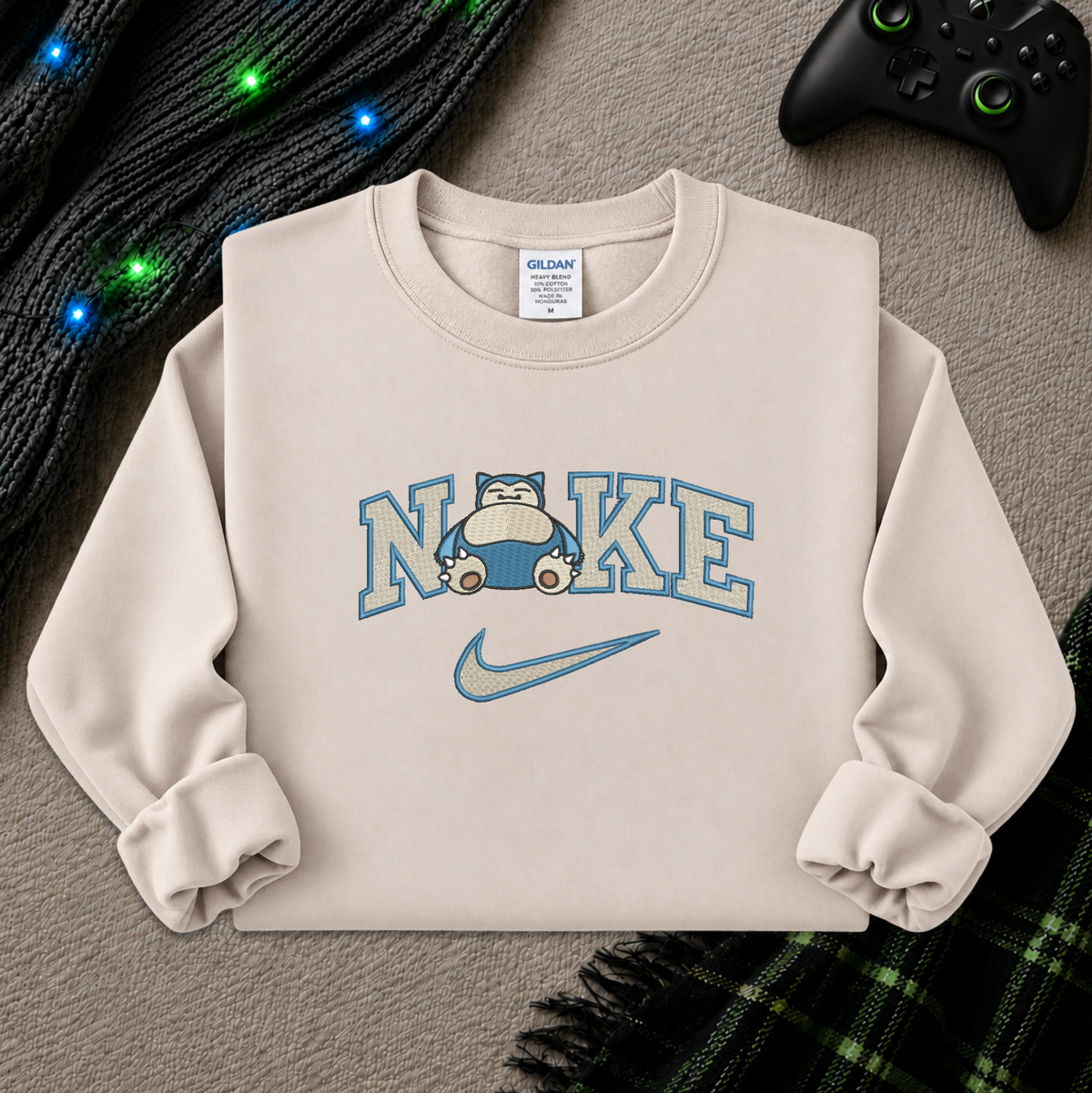 A beige crewneck sweatshirt featuring a 'NI KE' graphic with a cartoon pokemon snorlax character and a Nike swoosh, styled flat-lay on a textured beige surface with a game controller, knitted sweater, and plaid blanket.