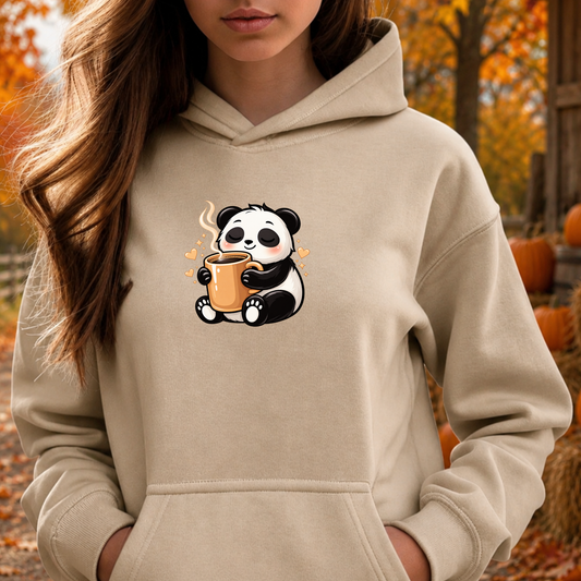 A woman wearing a beige hoodie with a cute cartoon panda holding an orange mug on the chest, photographed outdoors in an autumn setting with pumpkins and fall foliage.