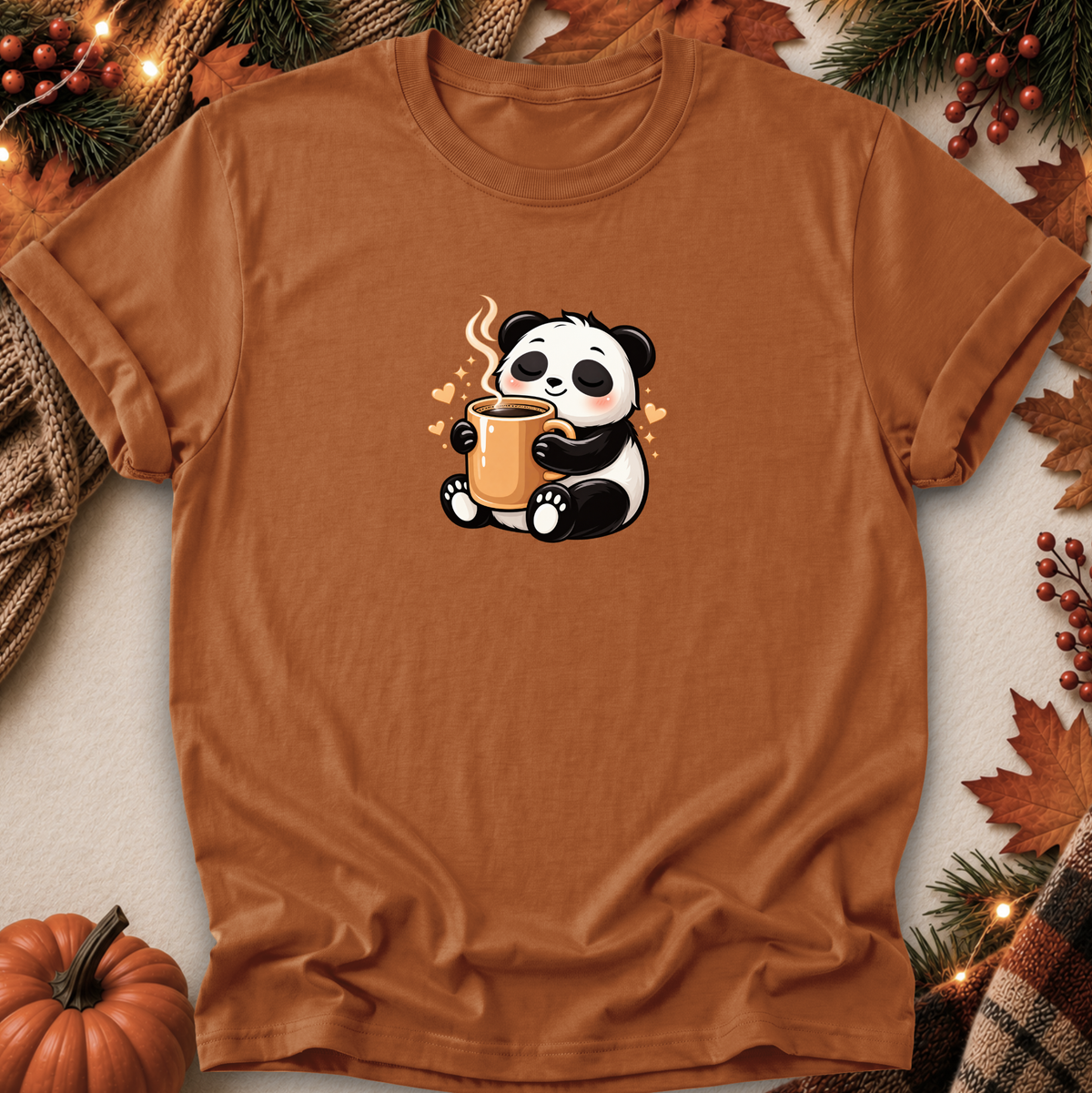 A rust-brown short-sleeve t-shirt with a cute cartoon panda holding a mug, styled in a flat-lay with autumn decorations like a pumpkin, pine branches, and leaves.