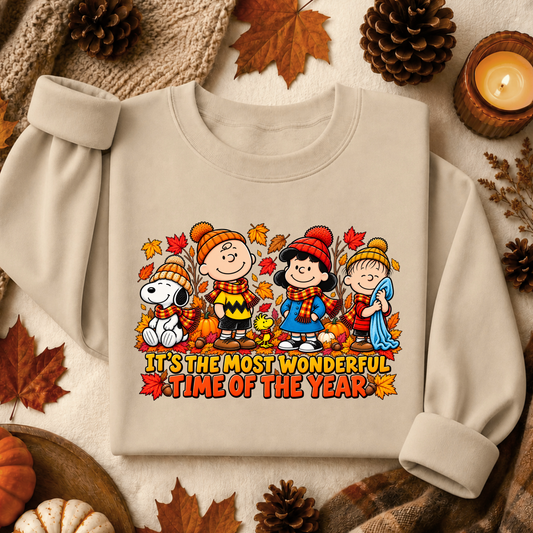 A beige crewneck sweatshirt featuring a colorful cartoon graphic of Peanuts characters in autumn clothing with the text 'IT'S THE MOST WONDERFUL TIME OF THE YEAR'. The sweatshirt is styled in a cozy flat-lay arrangement with autumn props including pinecones, maple leaves, a candle, and a knit blanket.