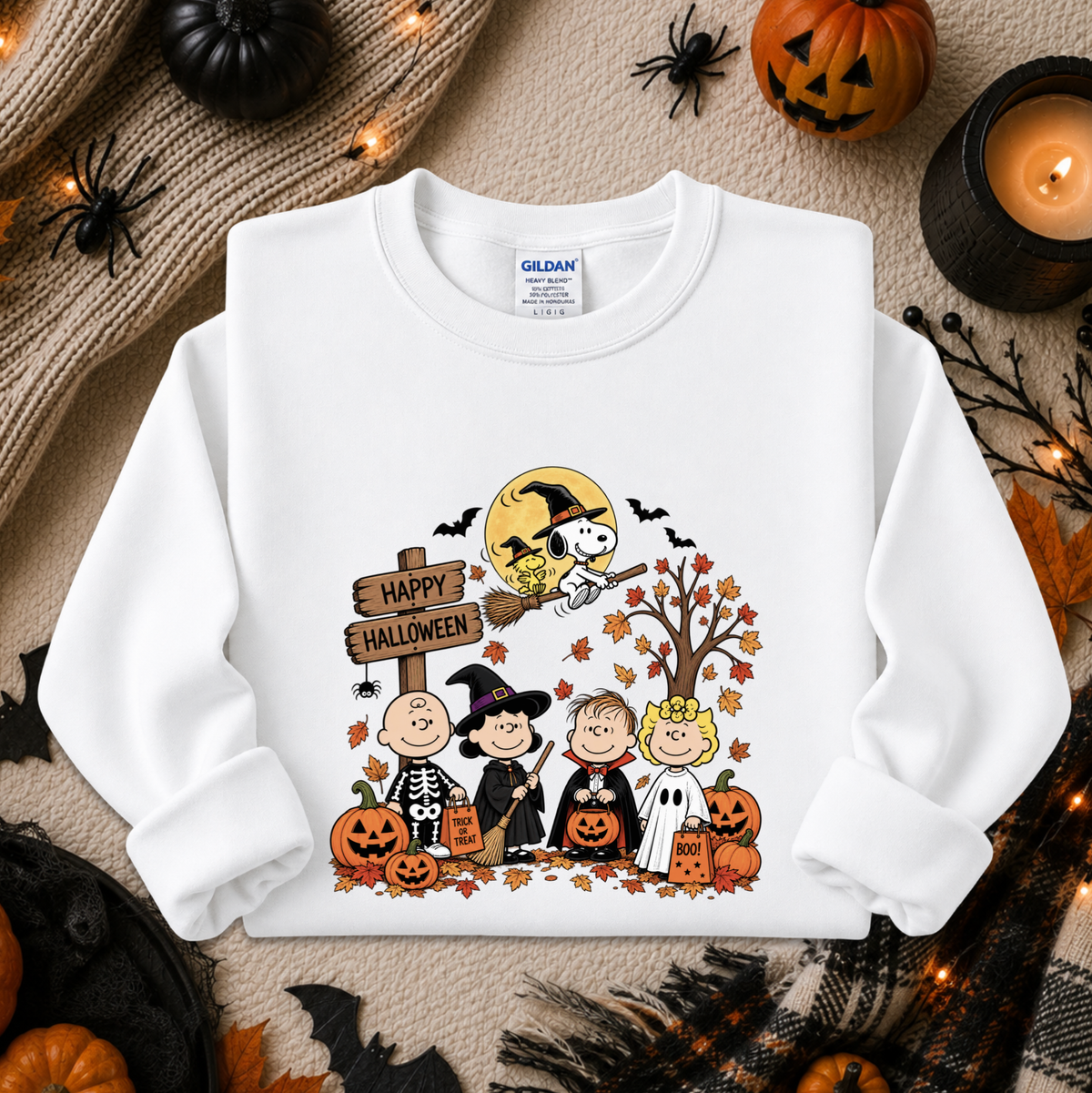 A white Gildan sweatshirt with a colorful 'Happy Halloween' cartoon graphic, styled in a flat-lay with seasonal props like pumpkins, a spider, and a candle.