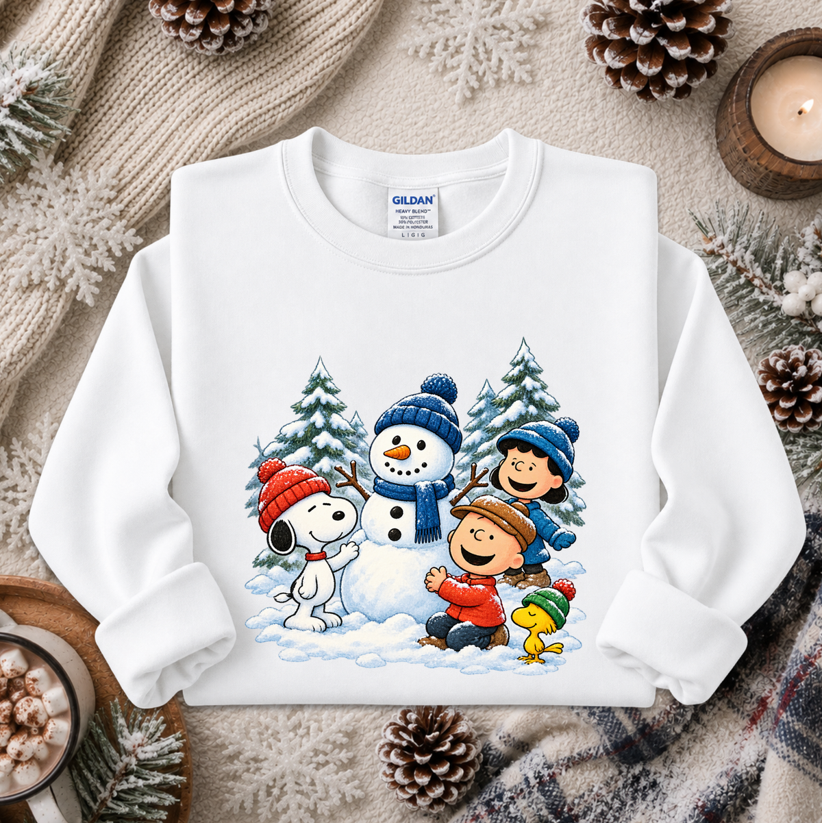A white long-sleeve crewneck sweatshirt featuring a colorful graphic of Peanuts characters Snoopy, Charlie Brown, Woodstock, and a snowman in a winter scene. The sweatshirt is displayed in a cozy flat-lay setting with a knitted scarf, pinecones, and a lit candle.