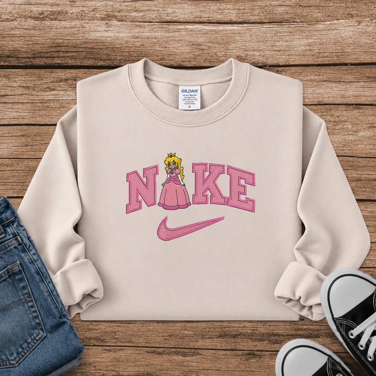 A beige crewneck sweatshirt styled in a flat lay on a wooden surface, featuring a pink 'Nike' graphic with a princess character and a pink swoosh. Blue jeans and black sneakers are partially visible as styling props.