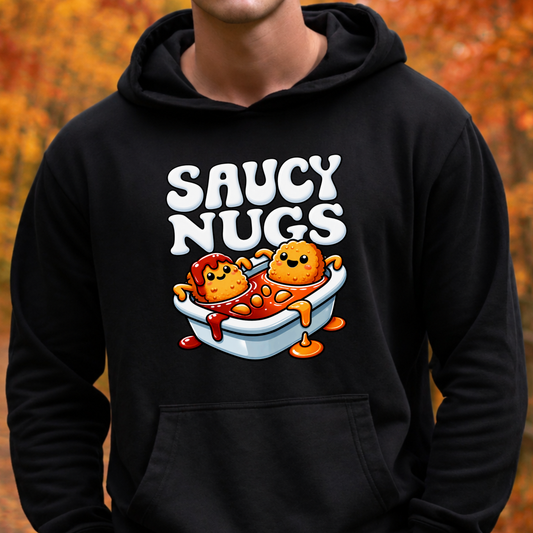 A person wearing a black pullover hoodie with a colorful graphic on the chest that reads 'SAUCY NUGS' above two cartoon chili dogs in a bathtub, set against an out-of-focus autumn background.