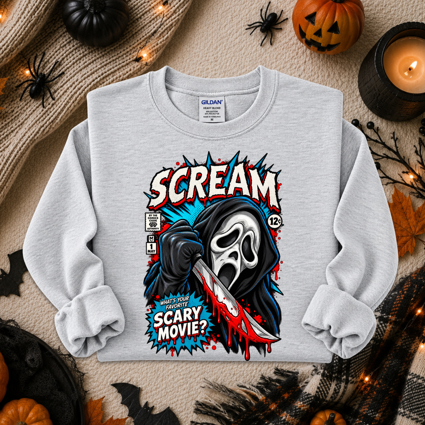 A light gray crewneck sweatshirt with a graphic print of Ghostface from Scream in a comic book style, featuring the word 'SCREAM' and a knife. The sweatshirt is displayed in a flat-lay arrangement with Halloween props including pumpkins, a candle, a spider, and a cozy knit blanket.