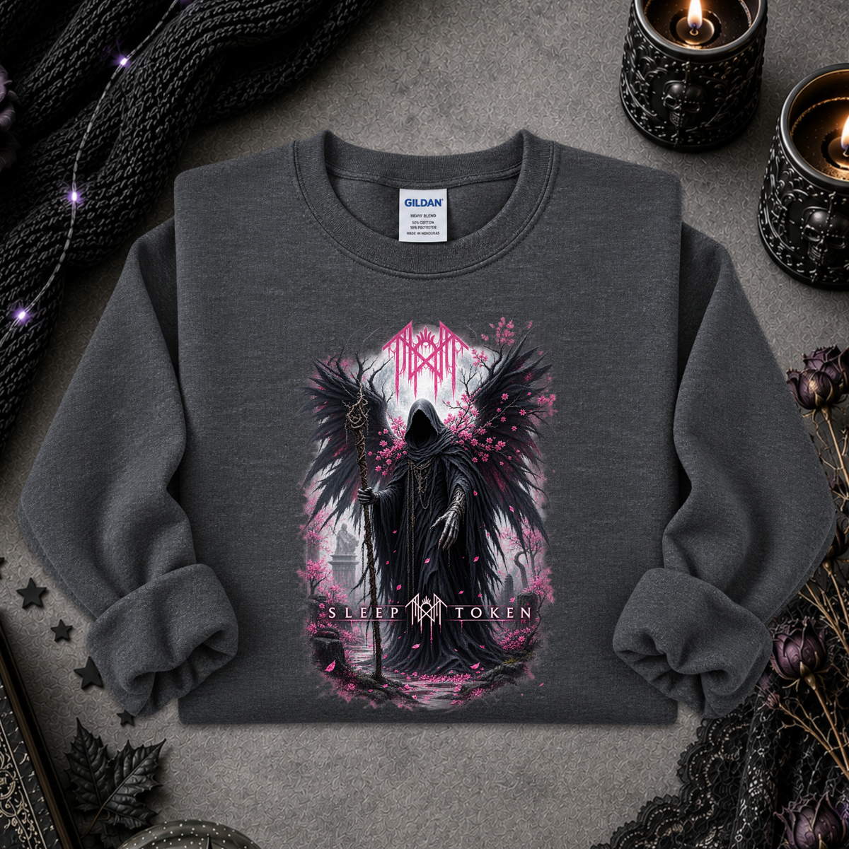 A dark gray crewneck sweatshirt with a gothic graphic of a winged hooded figure and pink blossoms, styled flat-lay on a textured gray surface with candles, lace, and black knit fabric.